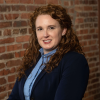 A professional headshot of a woman with fair skin and long, curly red hair. She is smiling slightly and wearing a navy blazer over a light blue button-up shirt. She has small stud earrings and is posed at a slight angle with her arms crossed. The background is a textured red brick wall, softly out of focus.